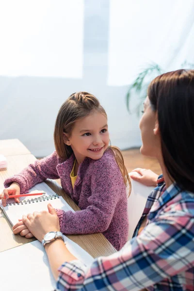 Happy daughter with pencil and notebook looking at mother while sitting at desk on blurred background — Stock Photo