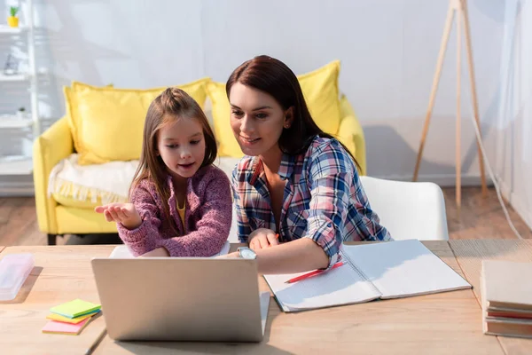 Sorrindo mãe olhando para laptop perto filha gesticulando na sala de estar — Fotografia de Stock