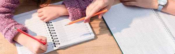 Vista recortada de la madre señalando con lápiz cerca de la hija escribiendo en el cuaderno en el escritorio, pancarta - foto de stock