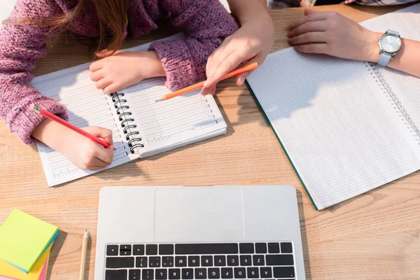 Cropped view of mother pointing with pencil near daughter writing in notebook near laptop on desk — Stock Photo