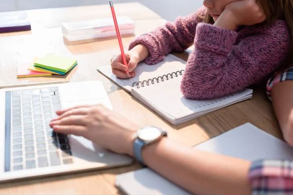 Vue recadrée de la fille écrivant dans un cahier au bureau avec une mère floue tapant sur un ordinateur portable au premier plan — Photo de stock