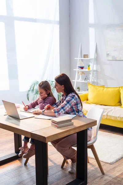 Full length of smiling mother sitting near daughter writing in notebook near laptop in living room — Stock Photo