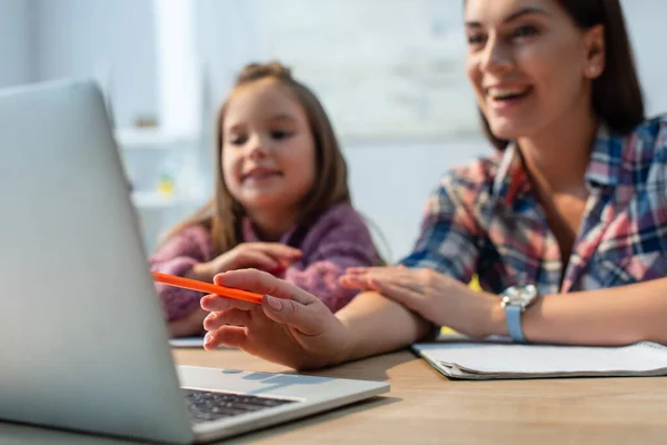 Усміхнена мати вказує олівцем на ноутбук біля дочки вдома на розмитому фоні — стокове фото