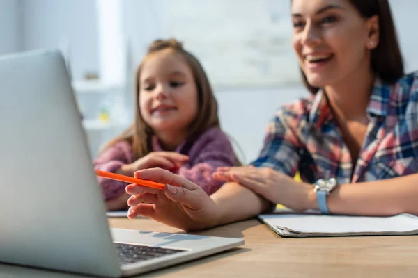 Mère souriante pointant avec un crayon à l'ordinateur portable près de la fille à la maison sur fond flou — Photo de stock