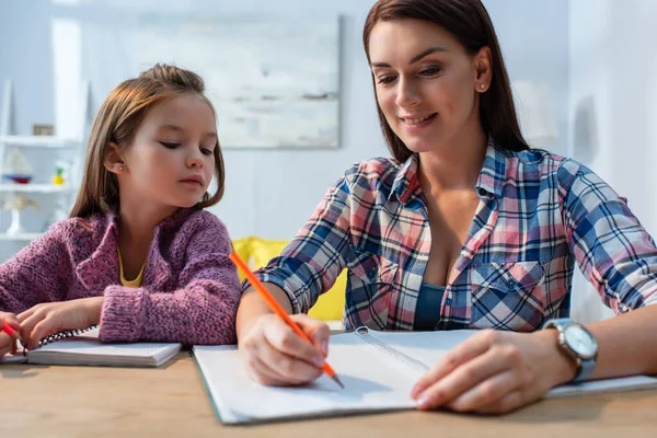 Sorridente madre scrivere in copia libro vicino figlia alla scrivania su sfondo sfocato — Foto stock