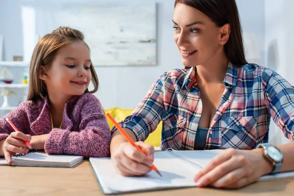 Smiling mother looking at daughter while writing in copy book at desk on blurred background — Stock Photo