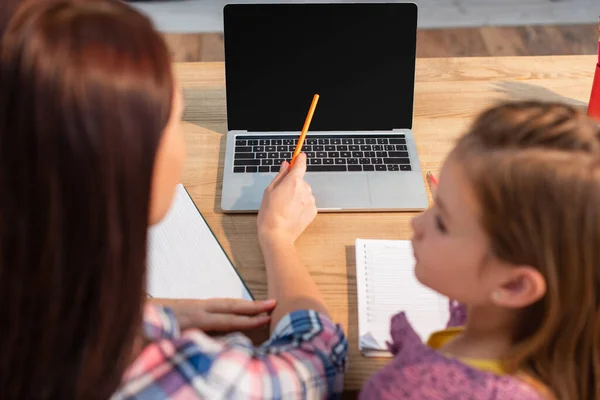 Hochwinkelaufnahme der Mutter, die mit Bleistift auf Laptop in der Nähe der Tochter zeigt, auf verschwommenem Vordergrund — Stockfoto