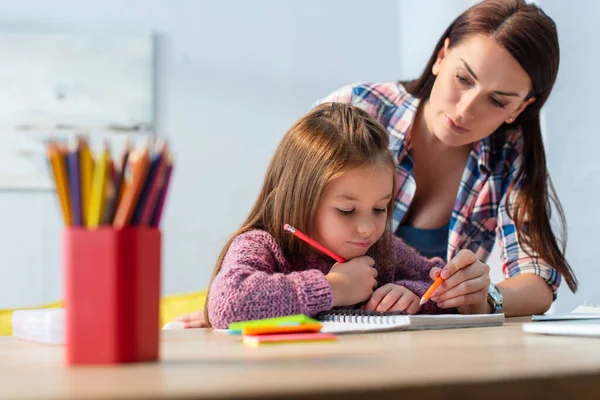 Mother pointing with pencil near positive daughter looking at notebook with blurred colored pencils on foreground — Stock Photo