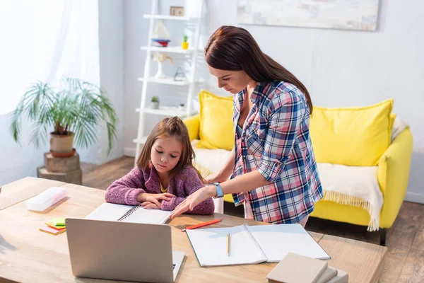 Madre seria señalando con la mano a la computadora portátil cerca de la hija en el escritorio sobre fondo borroso - foto de stock