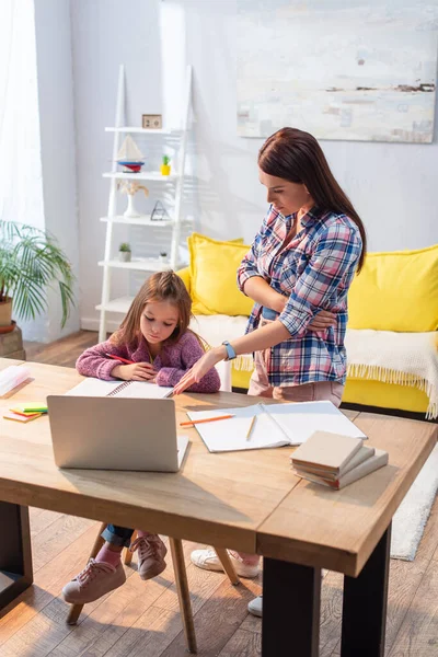 Full length of serious mother pointing with hand at laptop near upset daughter at desk in living room — Stock Photo