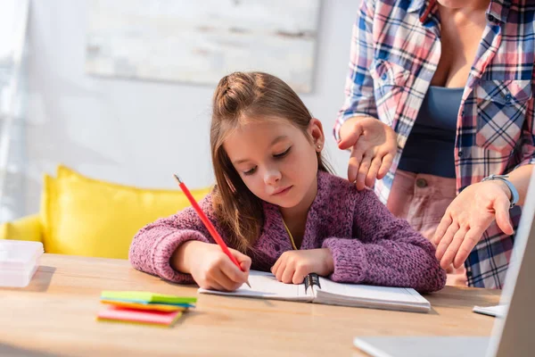 Mother gesturing near daughter writing in notebook at desk on blurred background — Stock Photo