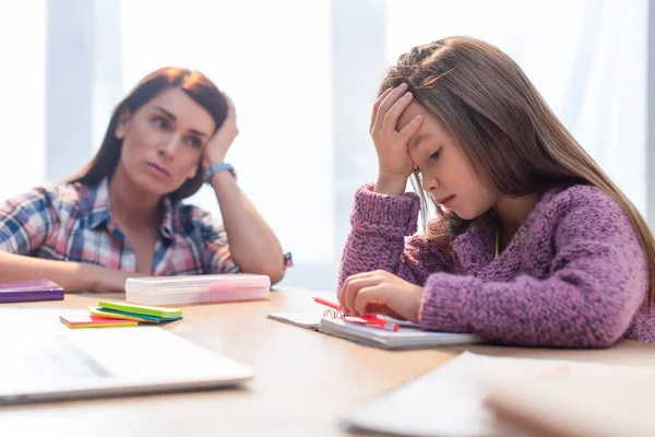 Menina cansada olhando para notebook na mesa com mãe preocupada borrada no fundo — Fotografia de Stock