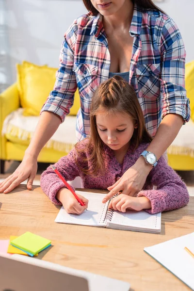 Мати вказує пальцем на блокнот, стоячи позаду дочки, пишучи за столом на розмитому передньому плані — стокове фото