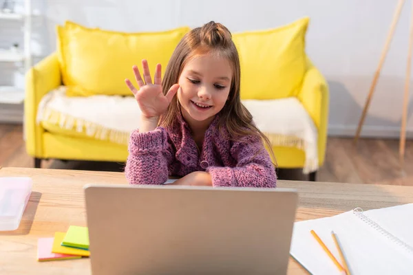 Lächelndes Mädchen mit winkender Hand und Blick auf Laptop auf Schreibtisch mit Papeterie im verschwommenen Vordergrund — Stockfoto