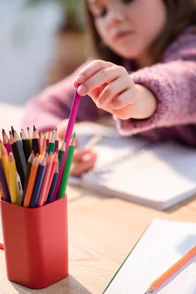 Обрізаний вид дівчини, яка бере кольоровий олівець з тримача на столі на розмитому фоні — стокове фото