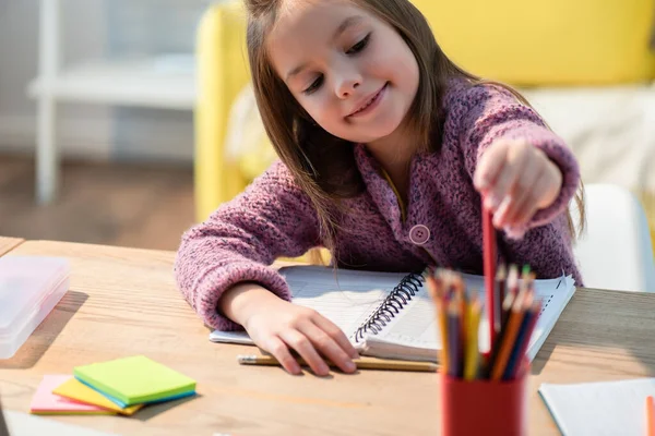 Улыбающаяся девушка берет цветной карандаш из держателя за столом с блокнотом и липкими заметками на размытом переднем плане — стоковое фото
