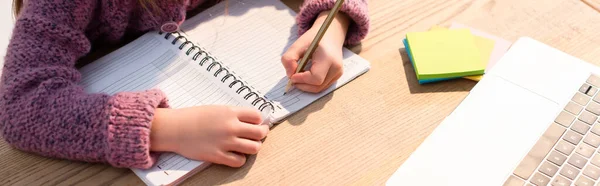 Обрізаний вид дівчини, що пише в блокноті біля липких нот і ноутбука на столі, банер — стокове фото