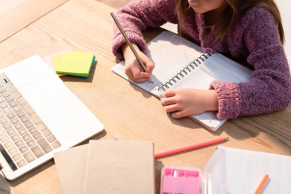 Vista cortada de menina escrevendo em notebook perto de notas pegajosas, livros e laptop na mesa — Fotografia de Stock