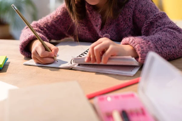 Vista recortada de la niña con la escritura a lápiz en el cuaderno en el escritorio en primer plano borroso - foto de stock