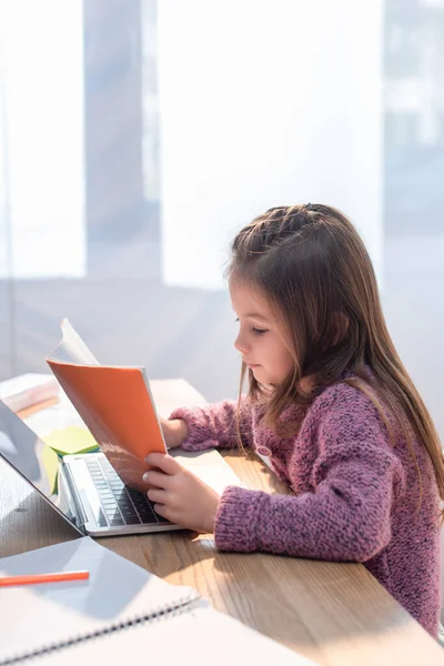 Fille regardant copie livre près ordinateur portable sur le bureau sur le premier plan flou — Photo de stock