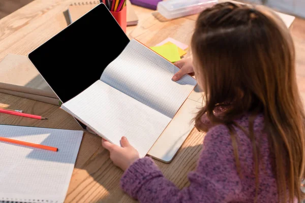 Vista de ángulo alto de niña sosteniendo libro de copias cerca de la computadora portátil con pantalla en blanco en el escritorio en primer plano borroso - foto de stock