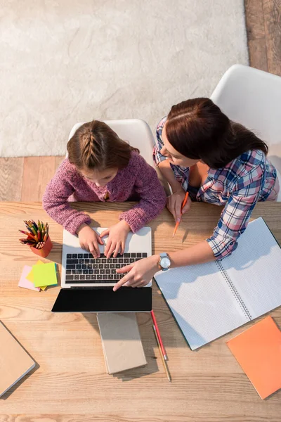 Visão superior da mãe apontando com o dedo para o laptop enquanto sentado perto da filha na mesa com artigos de papelaria — Fotografia de Stock