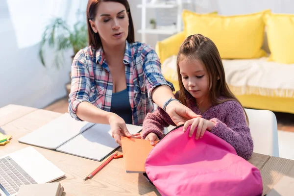 Mother and daughter putting copy book in backpack on desk at home on blurred background — Stock Photo