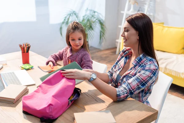 Happy mother putting copy book in backpack while sitting near daughter at desk on blurred background — Stock Photo