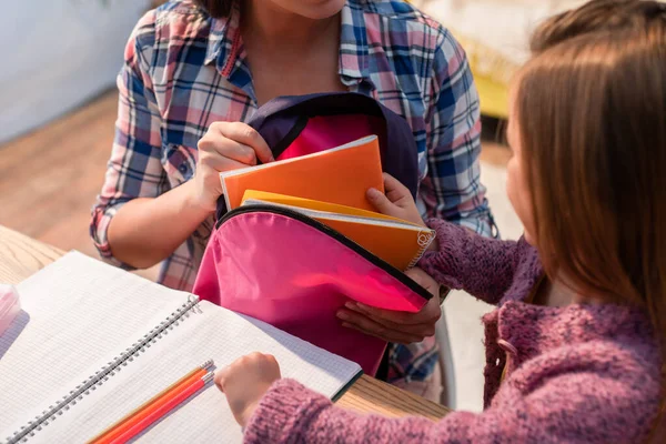 Tochter legt Kopierbuch in Rucksack neben Mutter und Schreibtisch auf verschwommenem Hintergrund — Stockfoto