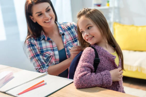 Happy mother putting backpack on daughter near desk at home on blurred background — Stock Photo