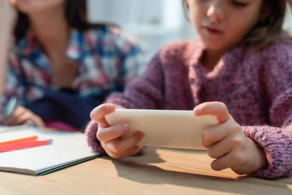 Vista recortada de la hija utilizando el teléfono inteligente cerca de la madre en el escritorio sobre fondo borroso - foto de stock