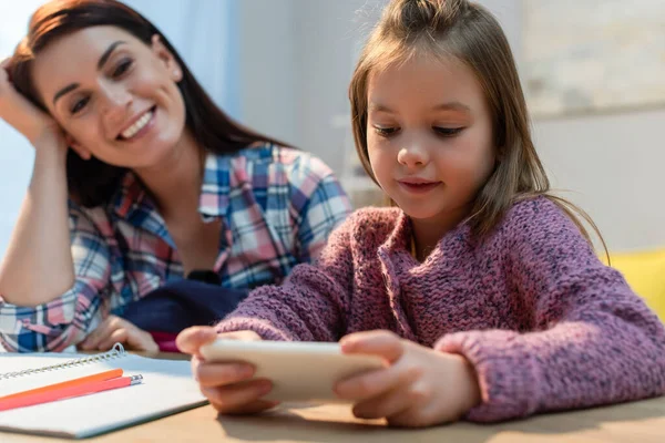 Positive daughter holding smartphone near mother at home on blurred foreground — Stock Photo