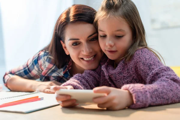 Happy mother and daughter with smartphone taking selfie at desk on blurred foreground — Stock Photo