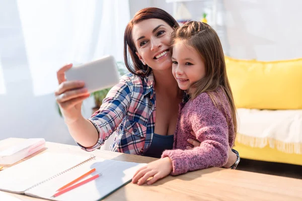 Fröhliche Mutter und Tochter machen Selfie am Schreibtisch mit Kopierbuch im unscharfen Vordergrund — Stockfoto