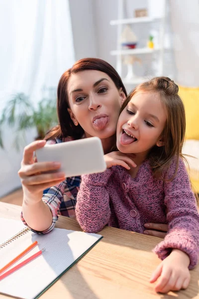 Mother and daughter with sticking out tongues taking selfie at desk on blurred foreground — Stock Photo