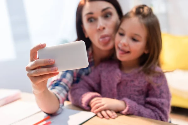 Madre e hija con lenguas que sobresalen tomando selfie en casa sobre fondo borroso - foto de stock