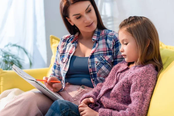 Madre con lápiz y libro de texto mirando a la hija en el sofá sobre fondo borroso - foto de stock