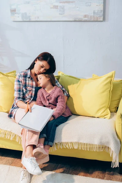 Full length of sad mother with copy book hugging daughter sleeping on couch at home — Stock Photo