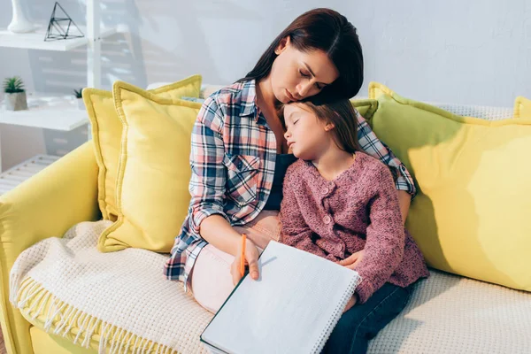 Madre molesta con lápiz y libro de copias abrazando a la hija durmiendo en el sofá - foto de stock