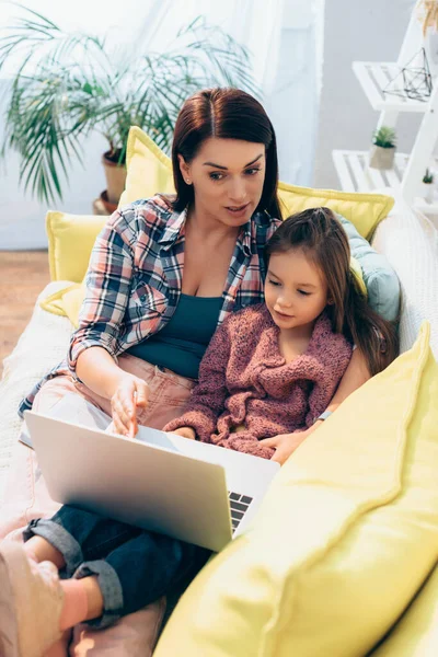 Mutter spricht und schaut auf Laptop, während sie Tochter auf Couch auf verschwommenem Hintergrund umarmt — Stockfoto