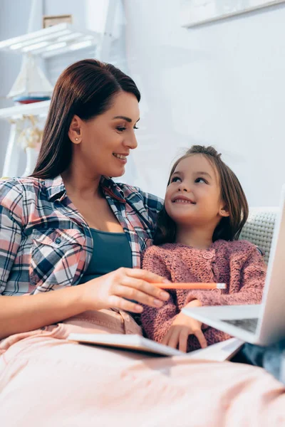 Smiling mother looking at daughter while pointing with pencil at blurred laptop on foreground — Stock Photo