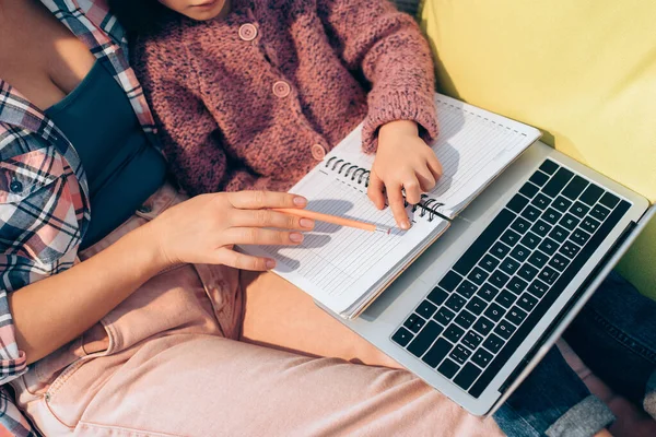 Ausgeschnittene Ansicht der Tochter, die mit dem Finger auf Notizbuch neben Mutter zeigt, mit Bleistift und Laptop auf Couch — Stockfoto