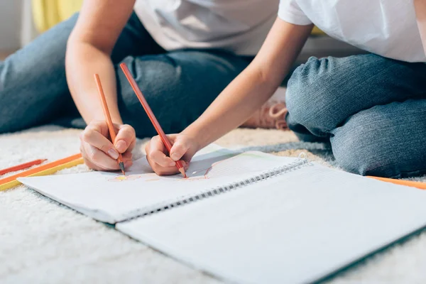 Cropped view of mother and daughter drawing with colored pencils in copy book on floor on blurred foreground — Stock Photo