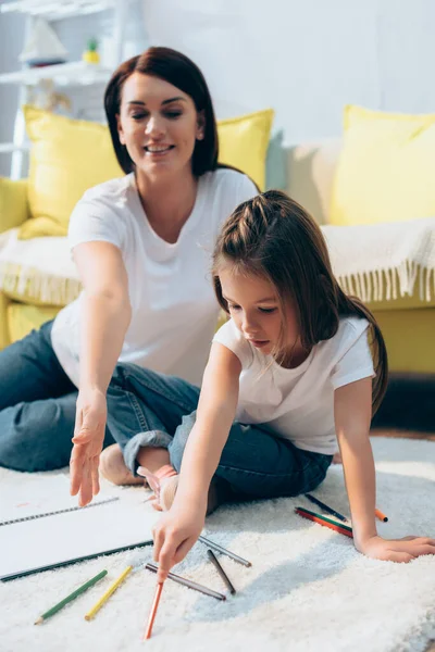 Daughter with colored pencil sitting near smiling mother on floor on blurred background — Stock Photo