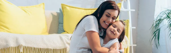 Madre feliz con los ojos cerrados abrazando hija cerca del sofá en casa, pancarta - foto de stock