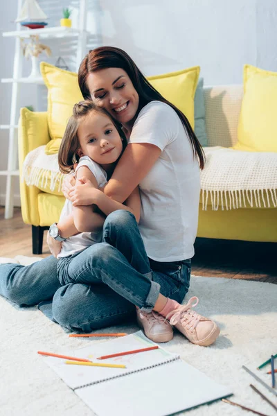 Cheerful mother with closed eyes hugging daughter while sitting on floor on blurred background — Stock Photo