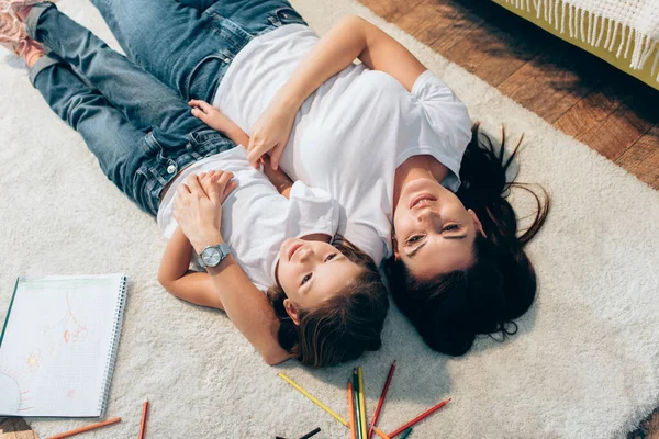 Overhead view of smiling mother and daughter hugging while lying on floor neat copy book and colored pencils — Stock Photo