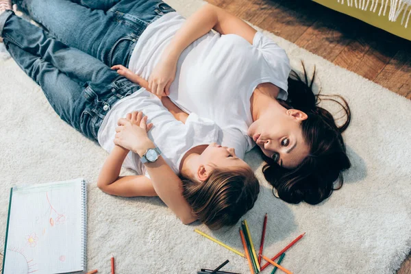 Vista aérea de la madre con los labios pucheros mirando a la hija mientras está acostada en el suelo cerca de la imagen y lápices de colores - foto de stock