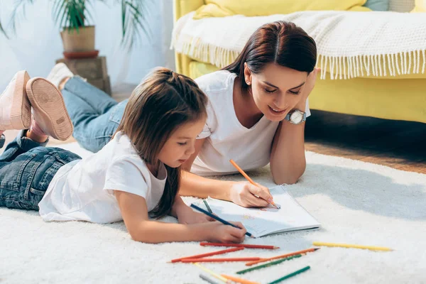 Positive mother and daughter with colored pencils drawing in copy book while lying on floor on blurred background — Stock Photo
