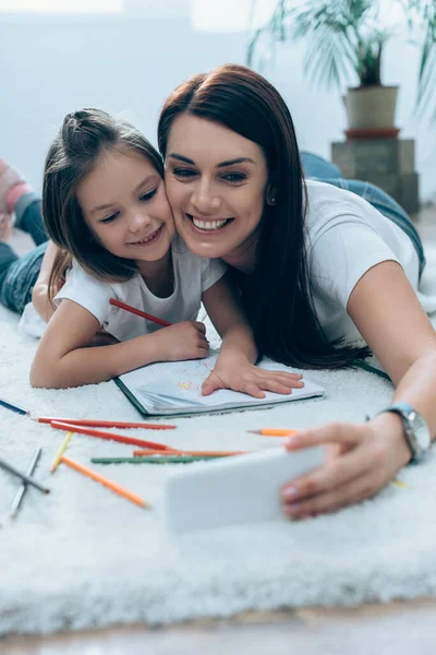 Усміхнена мати і дочка беруть селфі лежачи на підлозі біля кольорових олівців на розмитому передньому плані — стокове фото